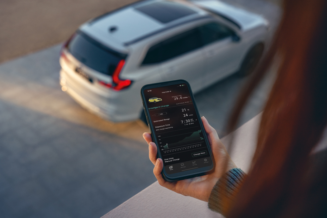Eine Hand hält ein Telefon, das die e:PROGRESS-App auf dem Bildschirm anzeigt, und auf der Straße.darunter parkt ein weißer Honda CR-V