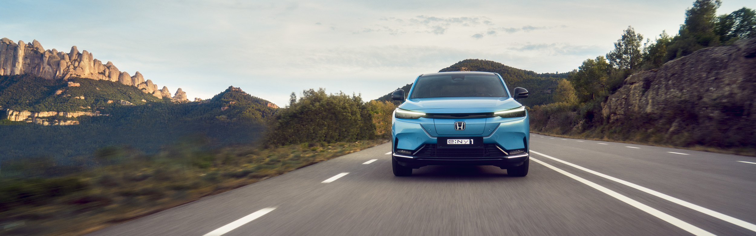 Frontansicht eines hellblauen Honda e:Ny1 auf einer Straße durch Hügel und Berge